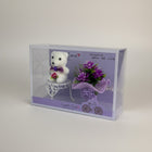 eRamis white plush teddy bear with purple bow and pink rose, sitting on a white table next to purple flowers.